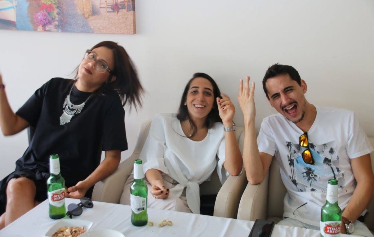 תמיר ושתי נשים צעירות יושבים ליד שולחן, צוחקים ומניפים ידיים.