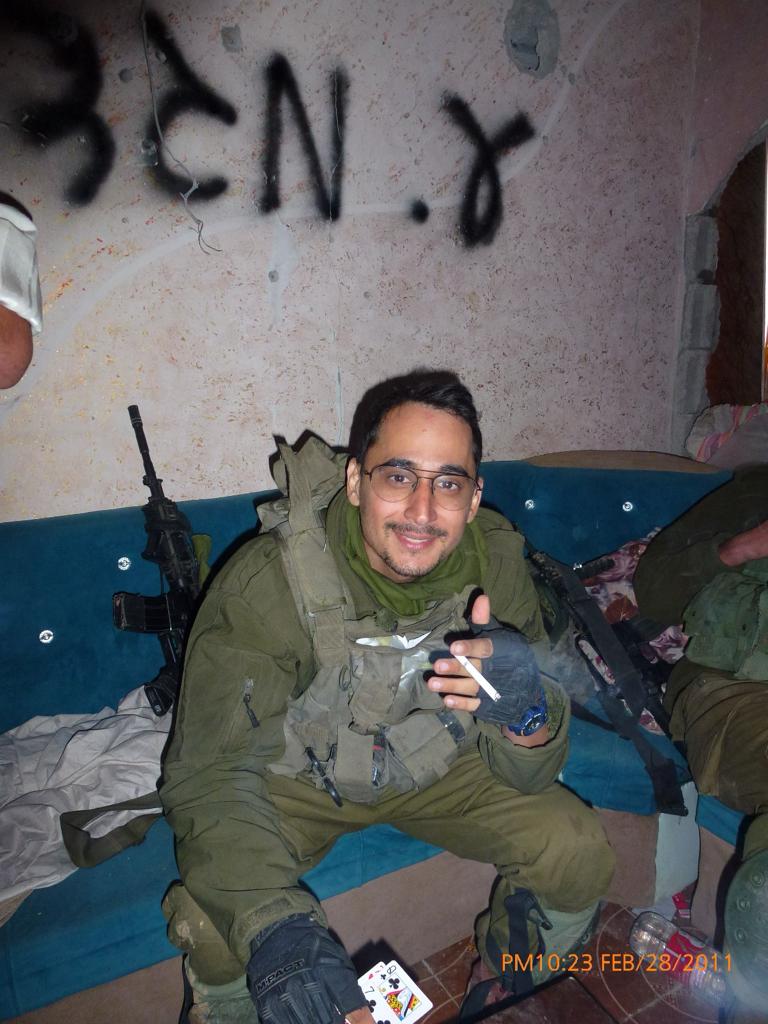 תמיר במדים ובציוד לחימה, יושב על ספה כחולה ומחייך למצלמה, קלפי משחק לרגליו.