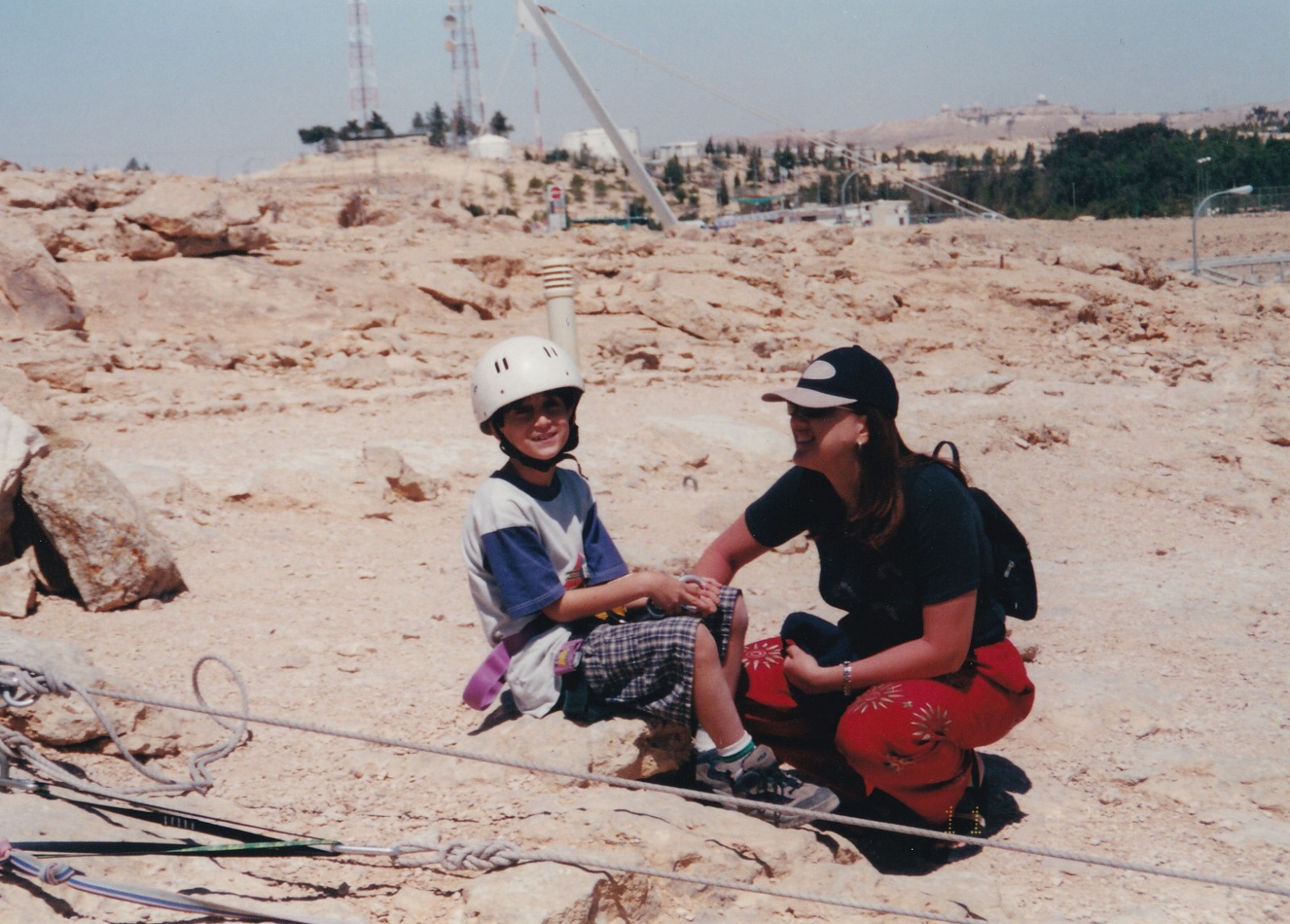 תמיר בילדותו חובש קסדה לבנה ויושב על סלע במדבר, ולצדו אישה בכובע מצחייה שמחייכת אליו.