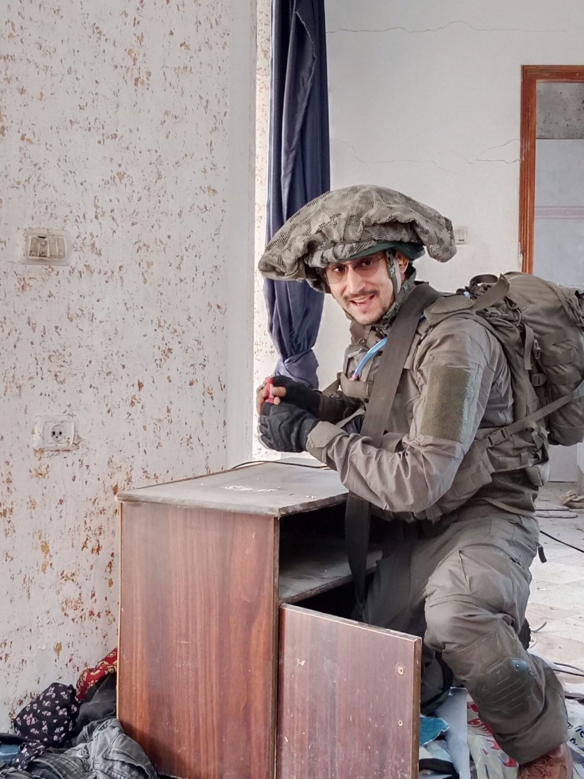 תמיר במדים, בקסדה ובתיק גב, כורע ליד ארונית עץ בחדר ומחייך למצלמה.
