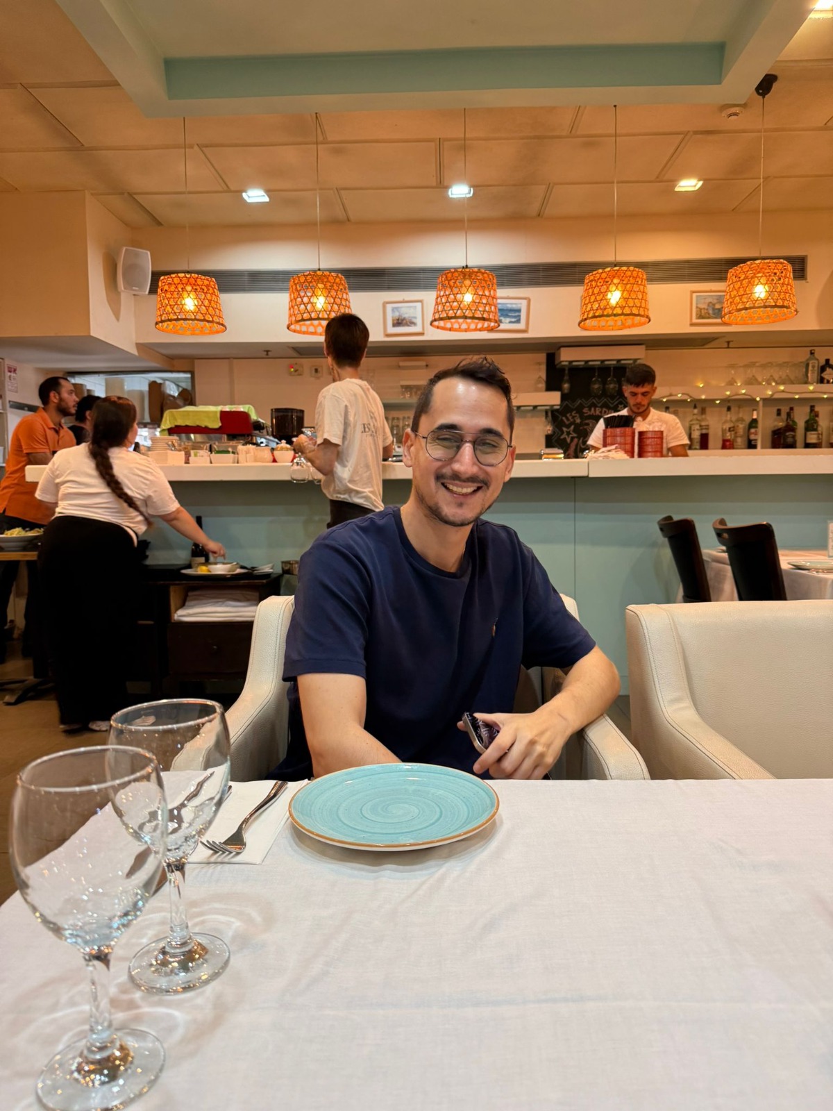 תמיר בחולצה כחולה ובמשקפיים יושב ליד שולחן במסעדה ומחייך חיוך רחב.