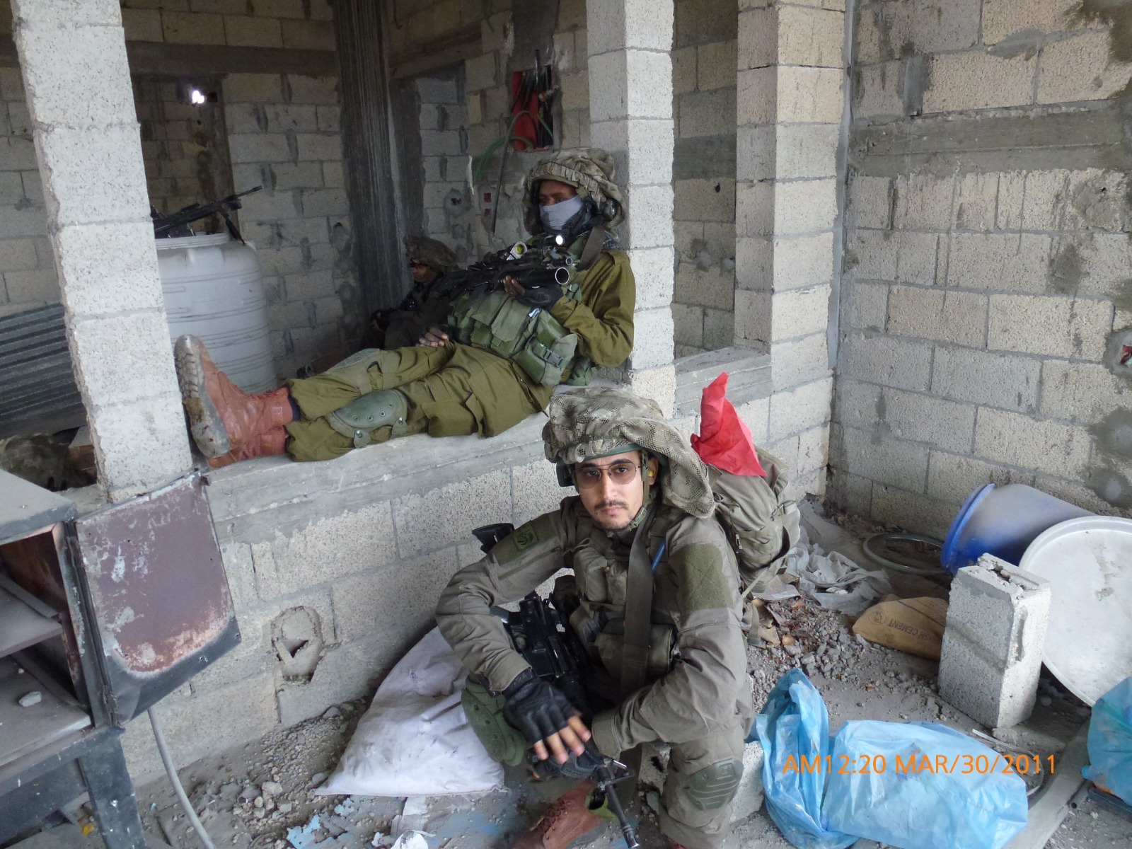 תמיר במדים ובקסדה יושב בין קירות בלוקים חשופים ומביט למצלמה, מאחוריו חיילים נחים.