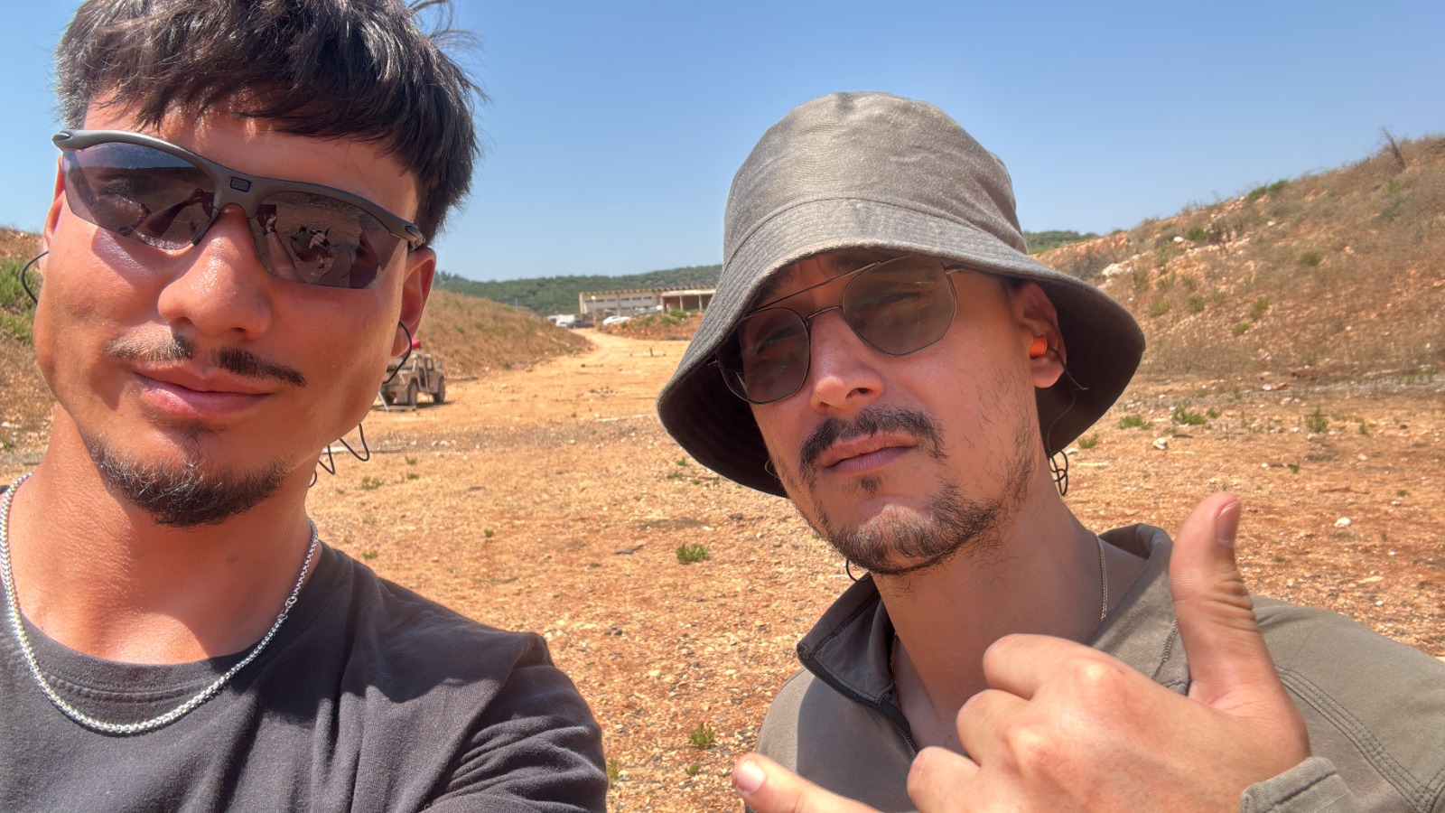 תמיר וחבר בסלפי בשטח פתוח ביום שמש, שניהם במשקפי שמש, אחד מהם מרים אגודל.