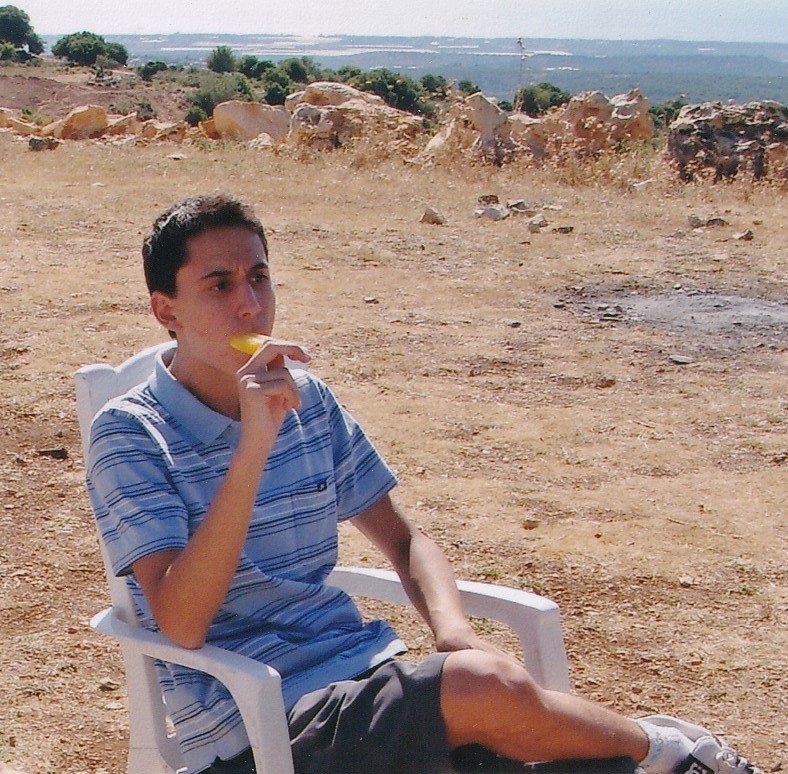 תמיר בנעוריו יושב על כיסא פלסטיק לבן בשטח פתוח ואוכל ארטיק.