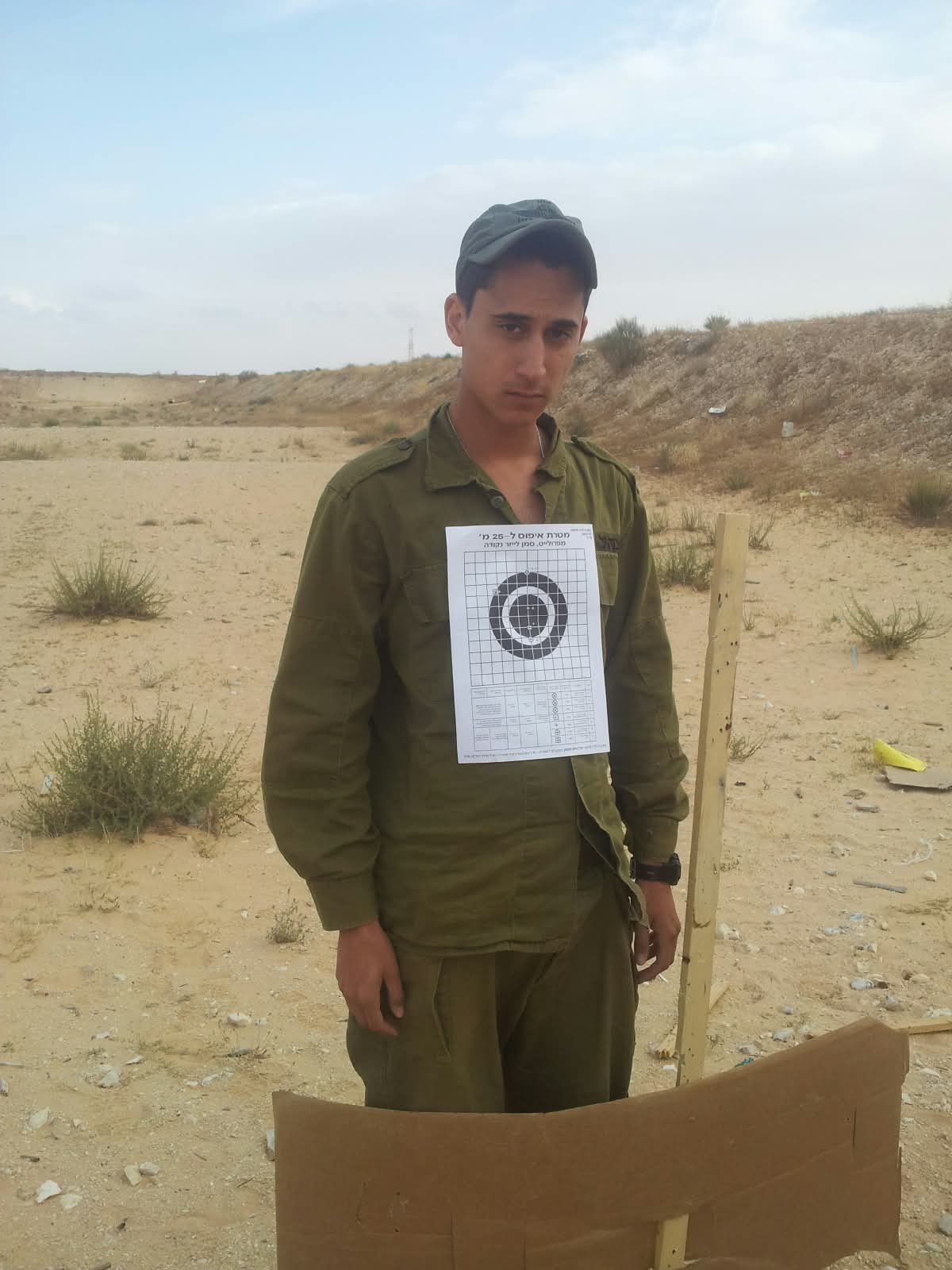 תמיר במדים ובכובע עומד במטווח, דף מטרה מוצמד לחולצתו.