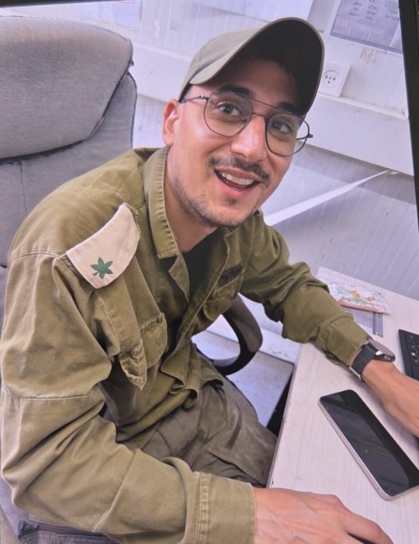 תמיר במדים, בכובע מצחייה ובמשקפיים, יושב ליד שולחן ומחייך למצלמה.