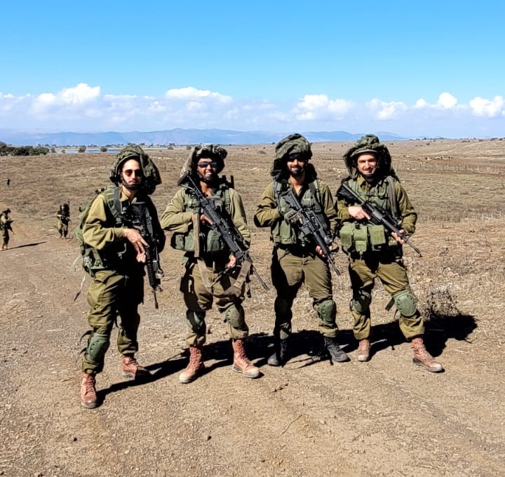 תמיר ושלושה חיילים נוספים בציוד מלא עומדים בשדה פתוח תחת שמיים כחולים.