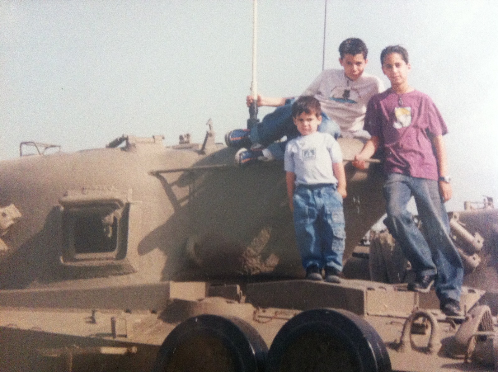 שלושה ילדים על טנק, שניים עומדים ואחד יושב מאחוריהם.