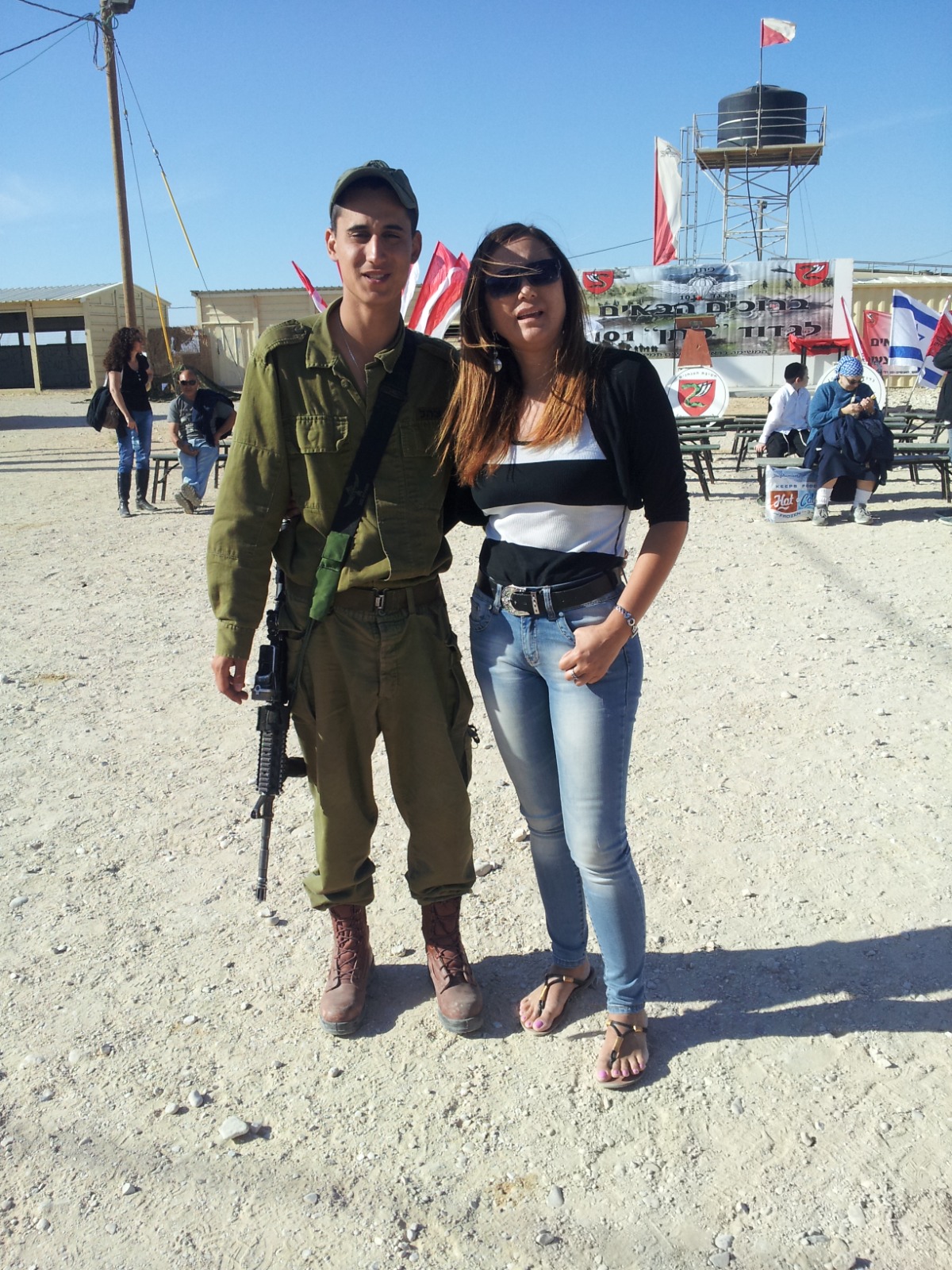תמיר במדים ובכובע, נשק על כתפו, עומד לצד אישה במשקפי שמש בטקס בבסיס. ברקע שלט ברוכים הבאים של גדוד 101.