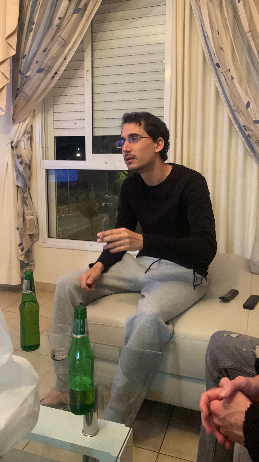 תמיר בחולצה שחורה ובמשקפיים יושב על ספה בסלון ומדבר, ידו מונפת באמצע משפט.