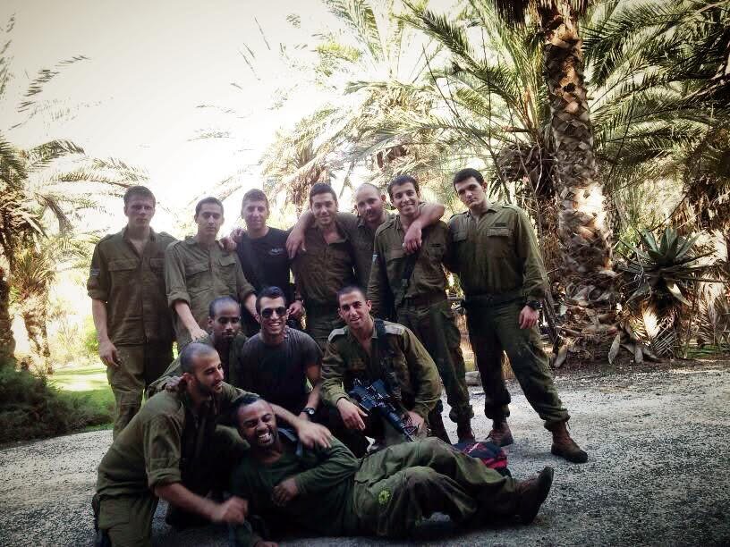 קבוצת חיילים מצטלמת יחד מתחת לעצי דקל, חלקם כורעים ואחד שוכב מלפנים וצוחק.