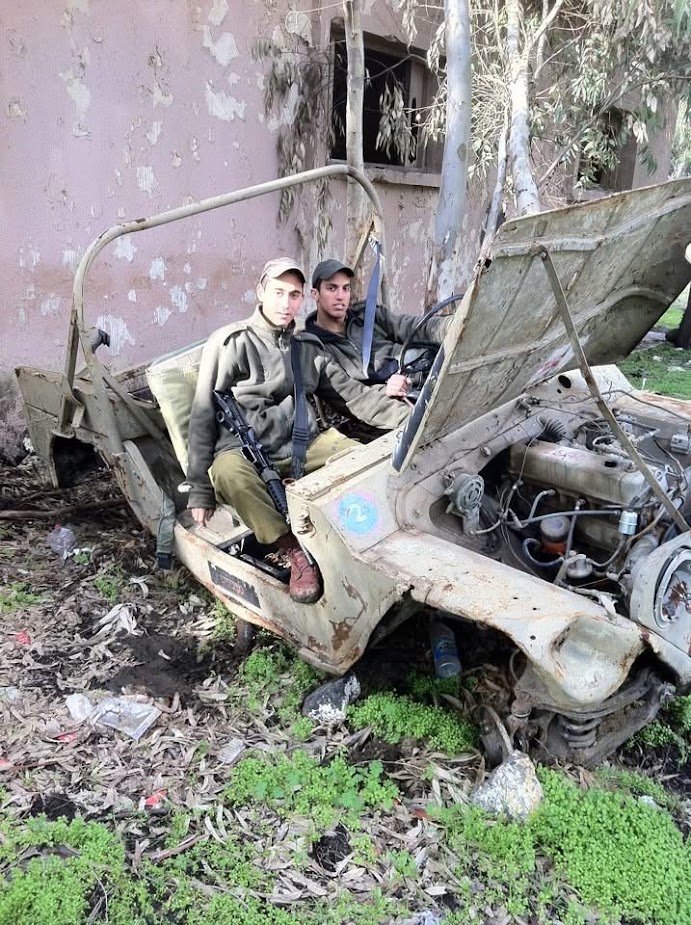 תמיר וחייל נוסף יושבים בג׳יפ ישן וחלוד בלי גלגלים, ליד בניין נטוש.