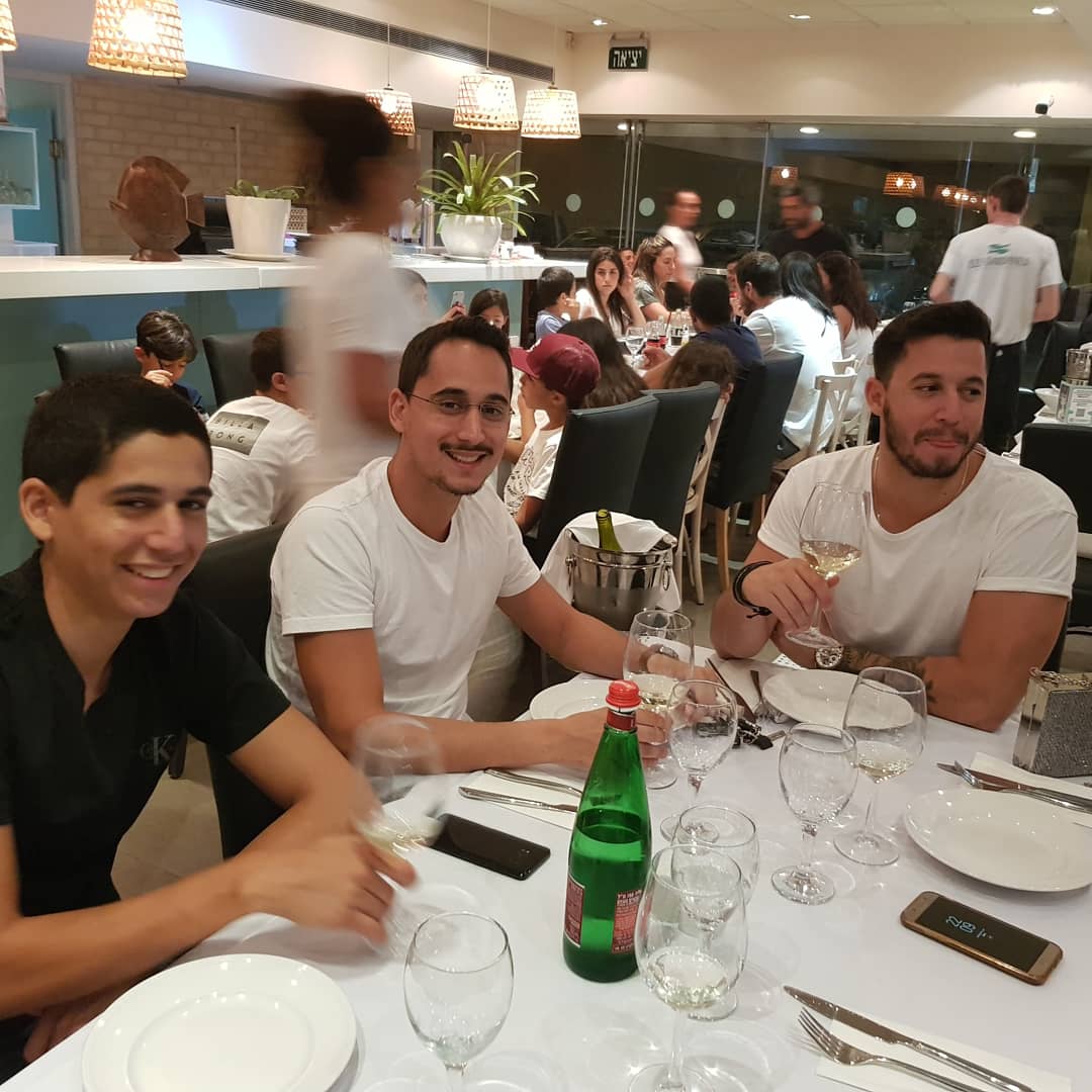 תמיר ושני צעירים נוספים יושבים ליד שולחן במסעדה ומחייכים, כוסות יין על השולחן.